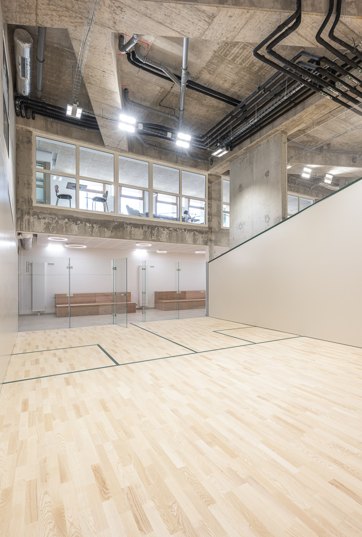 Terrain de squash double hauteur avec tribunes, mezzanine en hauteur sur rue et ambiance douce mais moderne avec béton brut technique apparente